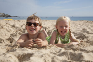 A boy and girl lying at Mission Beach, San Diego, on their stomachs on the sand, laughing and looking at the camera.