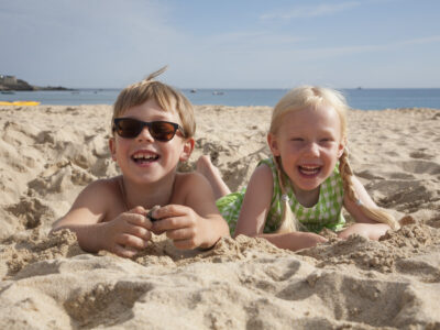 A boy and girl lying at Mission Beach, San Diego, on their stomachs on the sand, laughing and looking at the camera.