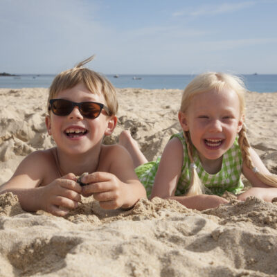 A boy and girl lying at Mission Beach, San Diego, on their stomachs on the sand, laughing and looking at the camera.